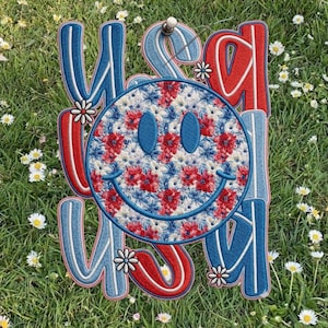 May include: Embroidered USA sign with a smiley face. The sign features red, white, and blue colors with a floral pattern on the smiley face. The sign is decorated with small white flowers and is placed on a bed of green grass.