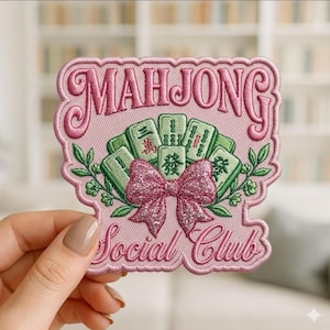 May include: Pink embroidered patch with the word "MAHJONG" at the top and "Social Club" at the bottom. The patch features green Mahjong tiles, a glittery pink bow, and green leaves.