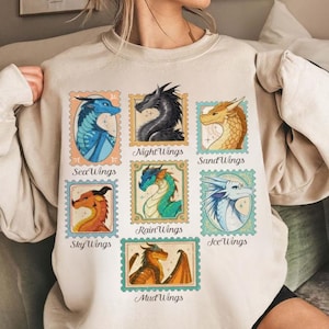 May include: A cream-coloured sweatshirt with a colourful graphic of dragon illustrations resembling postage stamps. Each stamp showcases a different dragon type, including SeaWings, NightWings, SandWings, SkyWings, RainWings, IceWings, and MudWings. Text labels each dragon type.