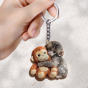 May include: A keychain featuring two monkey figures. One monkey is brown and hugging a gray monkey. The keychain has a silver chain and ring. The monkeys are made of a wood-like material.