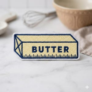 Puede incluir: Parche bordado con forma de barra de mantequilla. El parche es amarillo con un contorno azul y la palabra "BUTTER" en azul. Un diseño tipo regla está en la parte inferior. El parche es probablemente para ropa o accesorios.