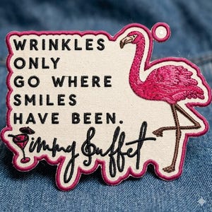 Puede incluir: Parche bordado con un flamenco rosa y la cita "Wrinkles Only Go Where Smiles Have Been". El parche también incluye el nombre "Jimmy Buffett" en cursiva, con una ilustración de cóctel.