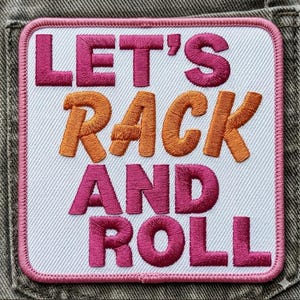 Puede incluir: Parche bordado con las palabras "LET'S RACK AND ROLL" en letras rosas y naranjas. El parche tiene un fondo blanco con un borde rosa y está cosido sobre una tela vaquera.