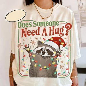 May include: Cream-colored t-shirt with a graphic of a raccoon wearing a Santa hat and string lights, with the text "Does Someone Need A Hug?" in red and green. The design includes snowflakes and a red and green border.