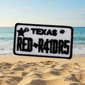 Puede incluir: Un parche rectangular con fondo blanco y texto y borde bordados en negro. El texto dice "TEXAS" encima de "RED-R41DR5". Una estrella y un corazón están a cada lado de "TEXAS". El parche está en una playa de arena con el océano al fondo.