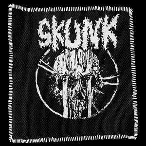 May include: Black square patch with white stitched border. The patch features the word "SKUNK" in a distressed font above a skull graphic within a circle. The skull has vertical lines resembling bars.