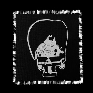May include: A black patch with a white, cartoonish illustration of a character with emo-style hair, sunglasses, and a skull and crossbones design. The patch has a white stitched border.