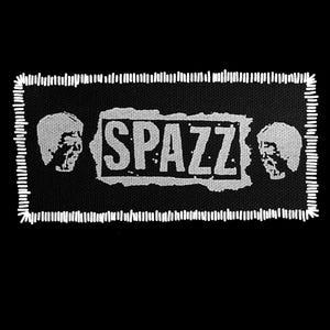 May include: Black rectangular patch with white stitched border. The word "SPAZZ" is printed in a white, distressed font inside a white rectangle. Two white, stylized faces flank the rectangle.