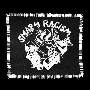 May include: Black square patch with white stitched border. The patch features the words "SMASH RACISM" in a stylized font, along with a graphic of a fist smashing through blocks. The design is in white against a black background.