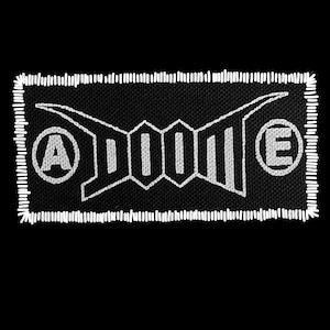 May include: Black rectangular patch with white stitched border. The patch features a white geometric design with the letters "A" and "E" in circles on either side of the central design. The word "DOOM" is in the center.