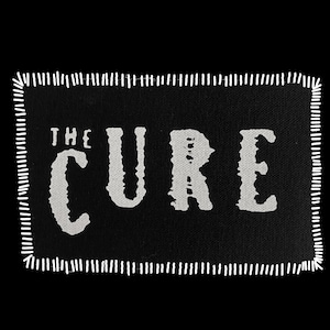 May include: A black rectangular patch with the words "THE CURE" in distressed white lettering. The patch has a white stitched border, creating a vintage or handmade aesthetic. The background is solid black.