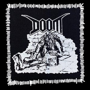 May include: Black and white patch with the word "DOOM" in a stylized font. The patch features a detailed illustration of a figure slumped over a box, set against a brick wall background. The patch has a white stitched border.