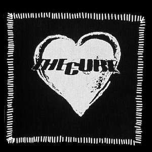 May include: Black square patch with a white heart design and the text "THE CURE" in a bold font. The heart is slightly distressed, and the patch has a white stitched border. The design is simple and graphic.
