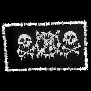 May include: A rectangular black fabric patch featuring white embroidered graphics of two skulls with dripping bone details and a central circle formed by bones crossed with two additional bones. The border has a thick white whip-stitch edge.