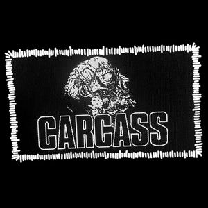 May include: Black rectangular patch with a white stitched border. The patch features a white illustration of a skull-like figure above the word "CARCASS" in bold, white capital letters. The design is set against a black background.