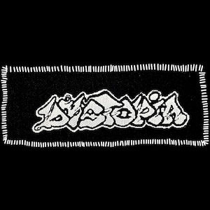 May include: Black rectangular patch with white stitched border. The patch features the word "SYNDROME" in a stylized, white, graffiti-style font. The patch is likely for clothing or accessories.