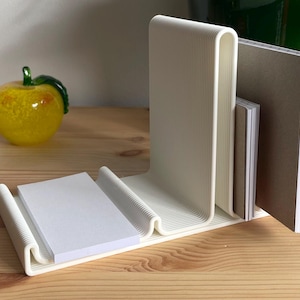 May include: A cream-coloured, modern desk organiser with a wave-like design. It holds a stack of white notepaper and several notebooks. A glass apple figurine sits to the left. The organiser is made of a ridged material.