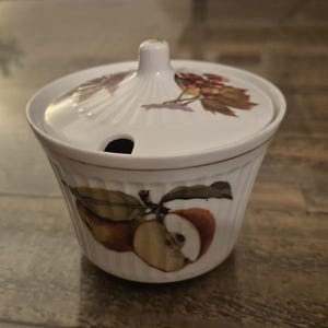 May include: White ceramic sugar bowl with a lid. The bowl is decorated with a fruit design featuring apples and berries in red, green, and brown. The lid has a small opening for a spoon. The bowl has a ribbed texture.