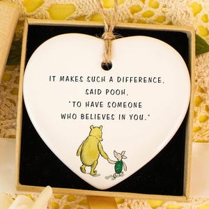 May include: A heart-shaped ceramic ornament with a quote from Winnie the Pooh, presented in a gift box. The quote reads, "It makes such a difference, said Pooh, 'to have someone who believes in you.'" A small gift box with a "Hand made" label and a decorative flower is also present.
