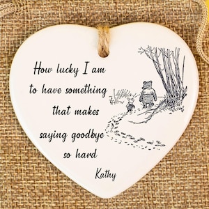 Może przedstawiać: Ceramiczna pamiątka w kształcie serca z ilustracją Kubusia Puchatka i cytatem "How lucky I am to have something that makes saying goodbye so hard Kathy." Umieszczona na worku z juty.