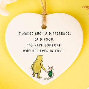 Puede incluir: Adorno de cerámica blanca en forma de corazón con una cita de Winnie the Pooh: "MARCA TAL DIFERENCIA, DIJO POOH, 'TENER A ALGUIEN QUE CREA EN TI." Presenta una ilustración de dibujos animados de Winnie the Pooh y Piglet.