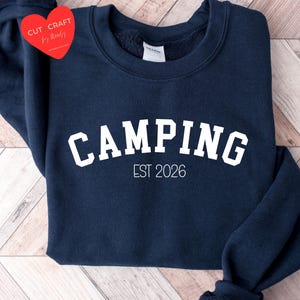 Könnte beinhalten: Ein marineblaues Sweatshirt mit dem Wort "CAMPING" in weißen, gebogenen Buchstaben. Unter dem Wort "CAMPING" steht der Text "EST 2026". Ein rotes herzförmiges Etikett befindet sich in der oberen linken Ecke.