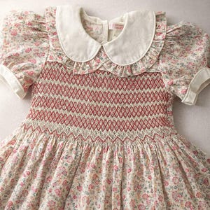 May include: A floral print dress with a white Peter Pan collar and short sleeves. The bodice features intricate red and white smocking. The dress has a full skirt and is made of a soft, lightweight fabric, suitable for warmer weather.