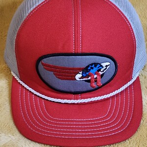 May include: Red and grey trucker cap with a white rope detail. The front panel is red with a grey patch featuring a winged design with red, white, and blue accents. The mesh back is grey.