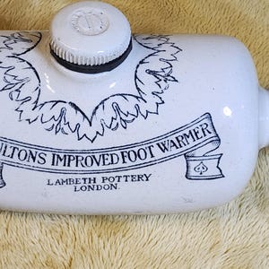 May include: Cream ceramic foot warmer with a screw-on cap. The body features a banner reading "DOULTON'S IMPROVED FOOT WARMER" and "LAMBETH POTTERY LONDON" in black script. Decorative floral elements surround the banner.