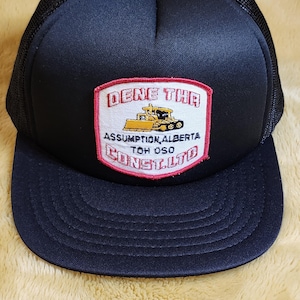 Peut inclure: Casquette de camionneur bleu marine avec un dos en filet noir et une visière incurvée. L'écusson avant affiche le texte "DENE THR" au-dessus d'un dessin jaune d'engin de chantier, avec "ASSUMPTION ALBERTA" et "TOH OSO CONSLTD" en dessous.