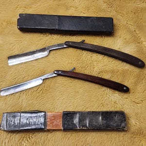 May include: Two straight razors with silver blades and dark wood handles are displayed. One handle is a lighter brown, the other is a darker brown. Two black cases are also present, suggesting a grooming or shaving set.
