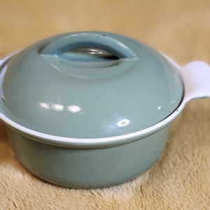 May include: A small, round, teal casserole dish with a matching lid and white handles. The dish has a white rim and a loop handle on the lid. The casserole dish is likely made of ceramic or enamel and is suitable for baking or serving.
