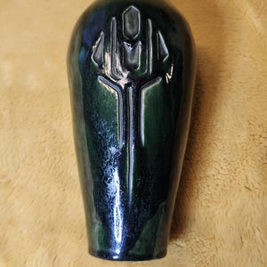Peut inclure: Un vase en céramique vert foncé avec une finition brillante et une forme cylindrique effilée. La surface avant présente un symbole en forme de trident stylisé, profondément gravé et centré verticalement, avec des nuances de glaçure.