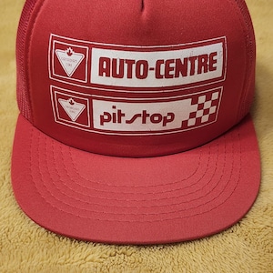 May include: Red trucker cap with a mesh back and a curved peak. The front panel displays the text "AUTO-CENTRE" and "pit stop" in white, alongside the Canadian Tire logo. A classic accessory for car enthusiasts.