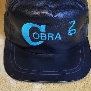 Peut inclure: Casquette de baseball en cuir noir avec le mot "COBRA" brodé en turquoise, ainsi que le chiffre "6". La casquette a une visière incurvée et un bouton sur le dessus. Le chapeau est posé sur une surface jaune.
