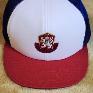 May include: A baseball cap with a red peak, white front panel, and blue mesh back. The cap has an embroidered emblem with a crest design. The crown is white, and the mesh is a dark blue.