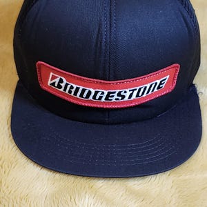 May include: Black baseball cap with a mesh back and a flat peak. The front has a red rectangular patch with the white "BRIDGESTONE" logo. The cap is likely made of a durable fabric, suitable for outdoor wear.