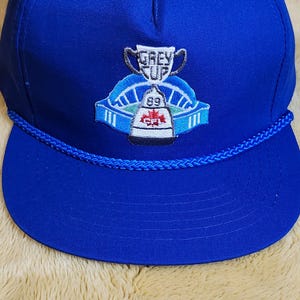 May include: A royal blue baseball cap with a flat peak and a rope detail. The cap has an embroidered emblem with the words "GREY CUP" above a trophy and the number "89".