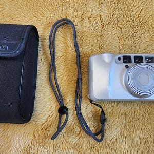 May include: A silver Minolta 110 Zoom camera with a black case and a grey neck strap. The camera has a lens, flash, and several buttons. The case has the Minolta logo.
