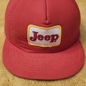 May include: A red baseball cap with a curved peak. The cap has a rectangular patch with the word "Jeep" in a serif font. The patch has a white background and a yellow border. The cap is made of a durable fabric.