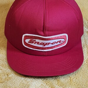 May include: A burgundy baseball cap with a curved peak. The cap displays the white and red "Snap-on" logo on the front. The hat is made of a durable material and has a classic six-panel design.