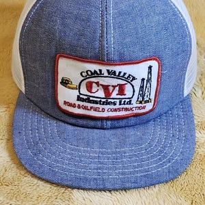 May include: A blue denim trucker cap with a white mesh back. The cap has a patch with the text "COAL VALLEY CVI Industries Ltd. ROAD & OILFIELD CONSTRUCTION" and a logo with a rig and a bulldozer.