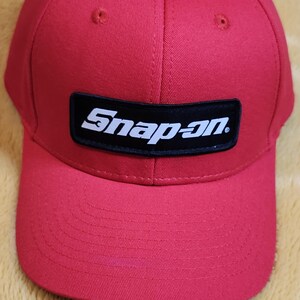 May include: A red baseball cap with a curved peak. The cap has a black rectangular patch on the front with the white text "Snap-on". The hat is made of a durable fabric and has a structured fit.