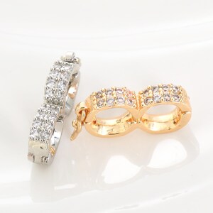 Puede incluir: Dos pares de pendientes. Uno es plateado, el otro dorado. Ambos presentan un diseño de doble aro, cada aro incrustado con pequeñas piedras preciosas transparentes. Los pendientes se muestran sobre una superficie blanca.