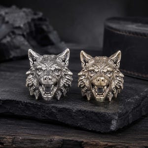 May include: Two rings featuring a detailed wolf head design with an open mouth showing teeth. One ring has a polished silver-tone finish, while the other has a muted, antique brass-tone finish, both showcasing intricate fur texture and facial features.