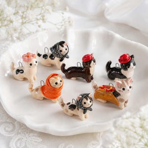Könnte beinhalten: Eine Sammlung von sieben kleinen, handbemalten Keramik-Katzenfiguren, die jeweils mit einem einzigartigen Accessoire wie einer Blume, einem Kopftuch, einem Barett oder einem Taco-Kostüm versehen sind. Jede Miniaturkatze hat eine kleine Metallöse am Rücken.