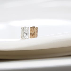 May include: Two rectangular metal beads with intricate embossed designs. One bead is silver, the other is gold. The beads are displayed on a white surface, showcasing their detailed patterns and metallic sheen. These beads are ideal for jewelry making.