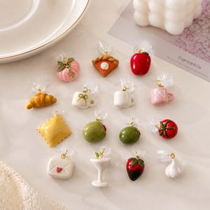 Könnte beinhalten: Eine Sammlung von Miniatur-Anhängern in Form von Lebensmitteln und Objekten, darunter ein rosa Kürbis, ein Stück Kürbiskuchen, ein roter Apfel, ein Croissant, Tassen, Ravioli, grüne Oliven, eine Tomate, ein Briefumschlag, ein Martiniglas, eine Erdbeere mit Schokolade und eine Knoblauchzehe.