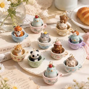 May include: A collection of small ceramic charms, each featuring a different animal sitting inside a miniature teacup. The animals include a fox, panda, hedgehog, cat, and birds, all equipped with a gold-toned metal loop for hanging as pendants.