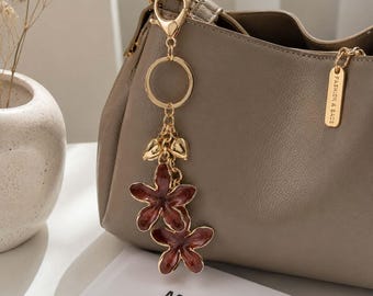 May include: A taupe leather handbag featuring a gold-toned metal keychain with two dark red enamel flower charms and small gold heart accents. A rectangular gold tag attached to the side of the bag is engraved with the text FASHION & BAGS.